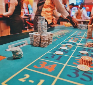 A casino scene with a roulette table features prominently, covered in green felt with colorful numbers and betting squares. Several stacks of poker chips are placed on different numbers, and the hands of several people are visible as they interact with the table. In the background, there are slot machines and more people, creating a vibrant and busy atmosphere.