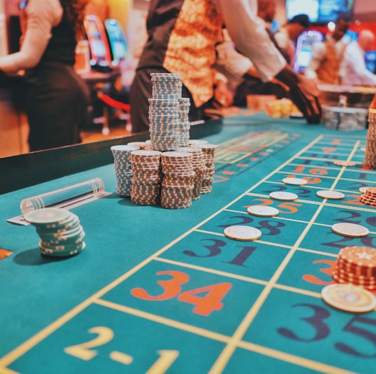A casino scene with a roulette table features prominently, covered in green felt with colorful numbers and betting squares. Several stacks of poker chips are placed on different numbers, and the hands of several people are visible as they interact with the table. In the background, there are slot machines and more people, creating a vibrant and busy atmosphere.