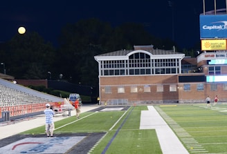 A nighttime scene featuring a sports stadium with a partially filled seating area and a large moon visible in the sky. The field is mostly empty except for a few people scattered around, and there are bright lights illuminating the venue. The stadium is surrounded by trees and a prominent sign displaying 'Maryland Lottery' is visible.