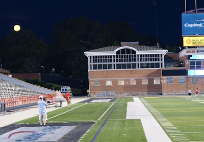A nighttime scene featuring a sports stadium with a partially filled seating area and a large moon visible in the sky. The field is mostly empty except for a few people scattered around, and there are bright lights illuminating the venue. The stadium is surrounded by trees and a prominent sign displaying 'Maryland Lottery' is visible.