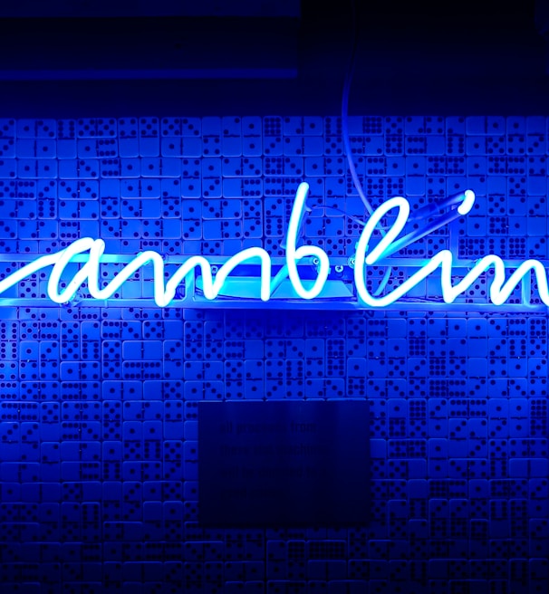 A bright blue neon sign spells out the word 'gambling' in cursive script. The background features a textured wall covered in domino tiles, also illuminated by the blue light, creating a vivid and visually striking scene.