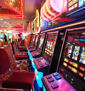 A brightly lit casino interior featuring a row of colorful slot machines. The neon lights create a vibrant atmosphere, with patterned carpeting adding to the decor. Stools are aligned neatly in front of each machine.