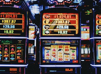 A row of colorful slot machines lit with bright neon lights, showcasing various graphical elements on the screens, indicating different games and jackpots. The atmosphere suggests a lively casino setting, bustling with excitement.