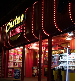 A brightly lit casino lounge entrance adorned with neon lights. The exterior features colorful signage including the words 'Casino' and 'Lounge' in vivid colors, with additional lights framing the building. Inside, a glimpse of a claw machine filled with plush toys and arcade games is visible. The area exudes an inviting yet bustling atmosphere typical of entertainment venues.