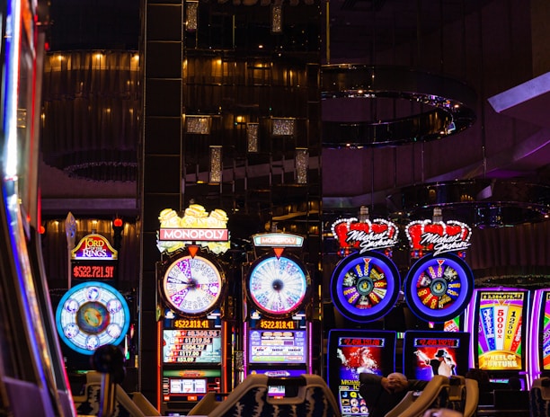 A lively casino scene featuring a row of bright, colorful slot machines with themes like Monopoly, Michael Jackson, and Lord of the Rings. The gaming machines display a variety of flashing lights and digital screens, creating an energetic and exciting atmosphere. Reflections of the lights are visible in glossy surfaces above the machines.