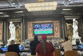 Several people are seated at a luxurious bar, with a large TV screen showing a sports game. The bar is elegantly decorated with ornate details and sculptures. The ceiling has a large, rectangular, crystal chandelier.