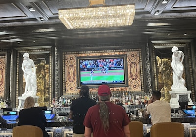 Several people are seated at a luxurious bar, with a large TV screen showing a sports game. The bar is elegantly decorated with ornate details and sculptures. The ceiling has a large, rectangular, crystal chandelier.