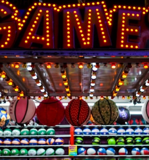 A brightly lit carnival game stall with a large 'GAMES' sign at the top surrounded by red and yellow lights. Various sports balls, including basketballs and soccer balls, are hanging from the ceiling and neatly arranged on shelves below.